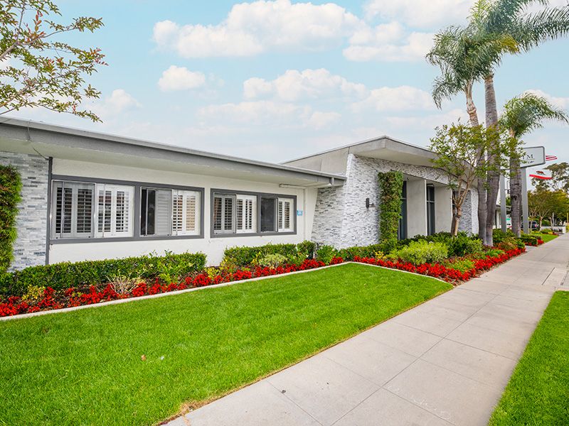 A single-story house with a well-maintained lawn, flower beds, and palm trees under a partly cloudy sky.