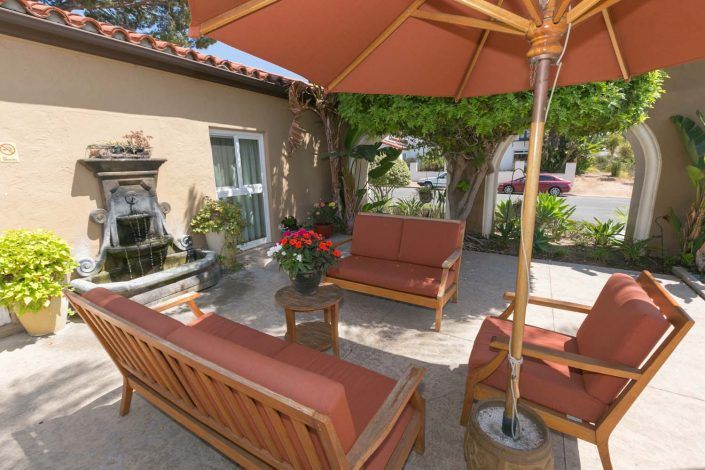A cozy patio setup featuring wooden outdoor furniture with red cushions and a large umbrella amidst garden greenery.