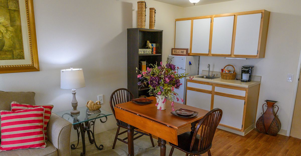 A cozy dining area with a wooden table set for two, adorned with a vase of flowers, adjacent to a kitchenette.