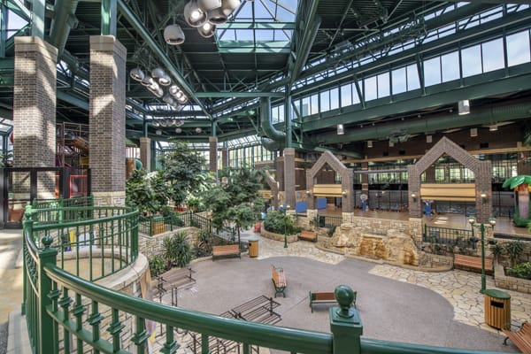 A spacious and well-lit atrium featuring industrial-style architecture with green metal beams, indoor plants, and wooden structures.