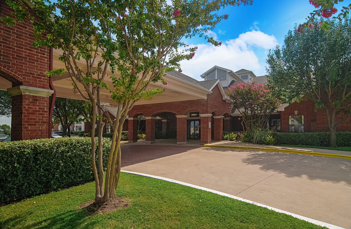 A brick building with a covered entrance surrounded by greenery and trees under a clear blue sky.