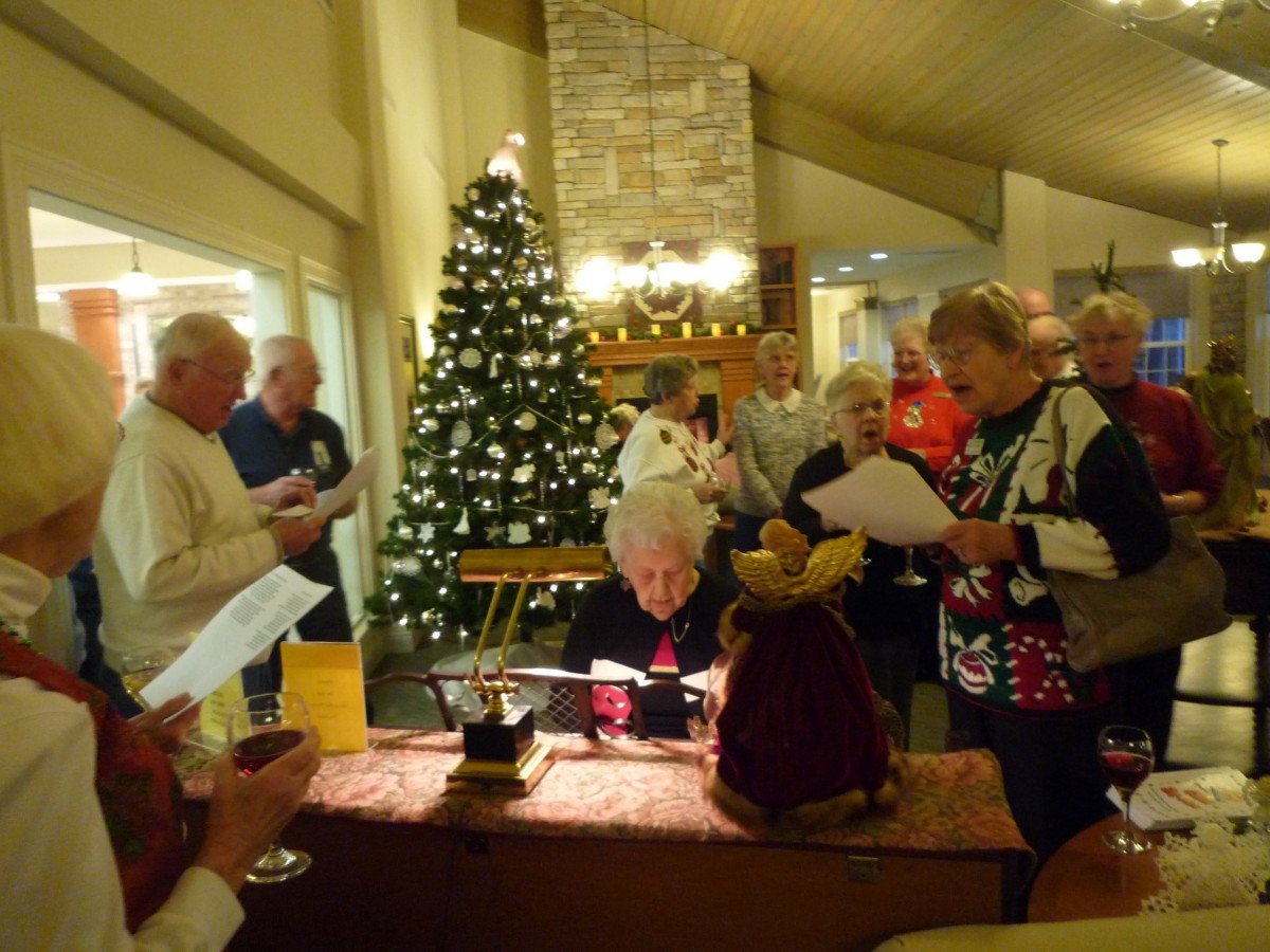 A group of people singing at a Christmas gathering, with a decorated tree and festive atmosphere.