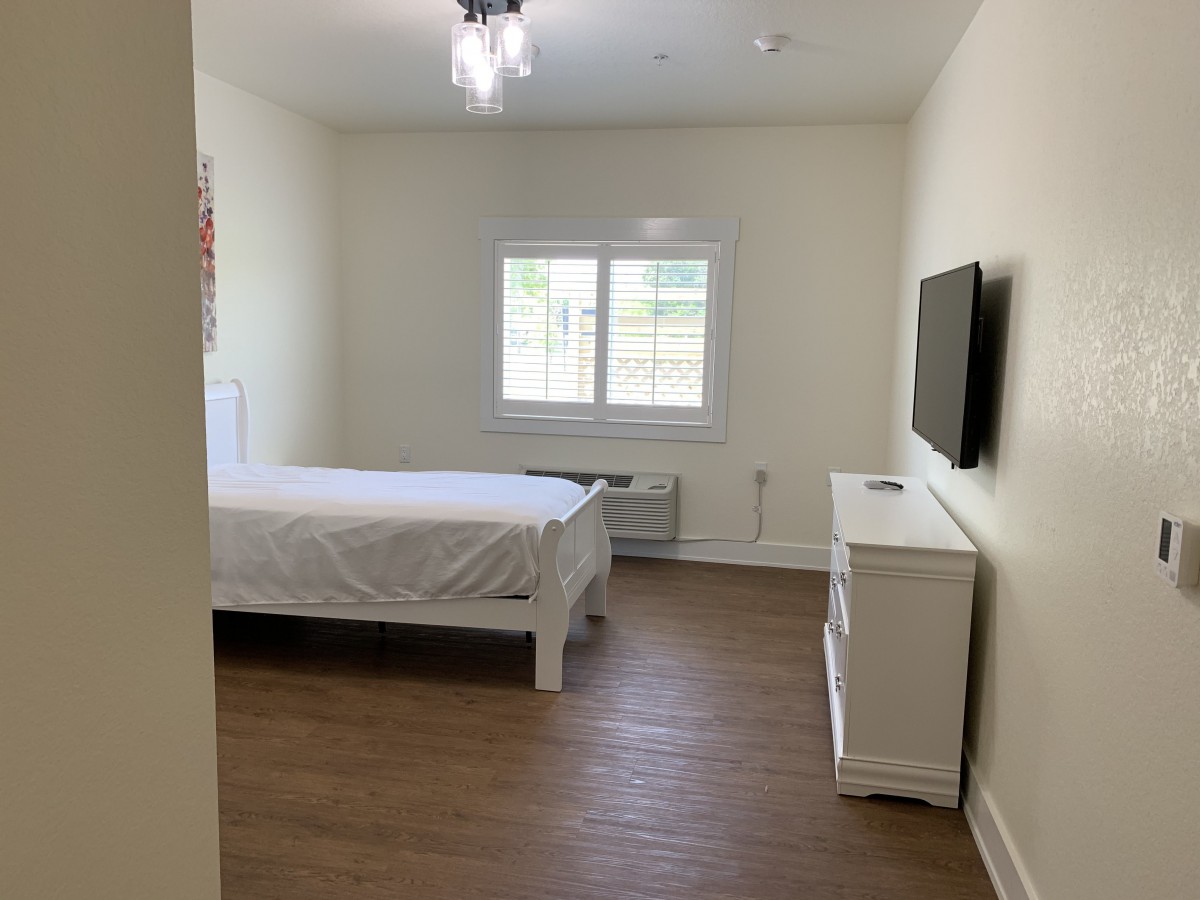 A minimalist bedroom featuring a bed, dresser, and wall-mounted TV, with natural light streaming through the window.