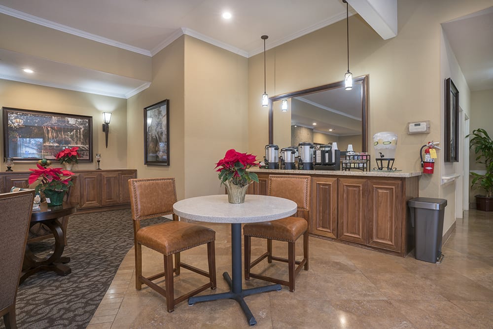A cozy dining area with a round table, two chairs, and a vase of red flowers, complemented by warm lighting and a mirrored cabinet in the background.