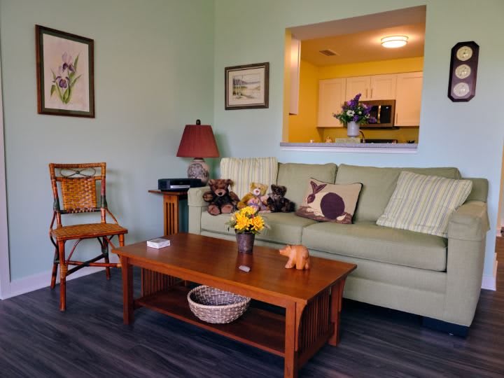 A cozy living room with a green sofa and wooden coffee table, featuring decorative cushions, artwork on the walls, and a view into the kitchen.
