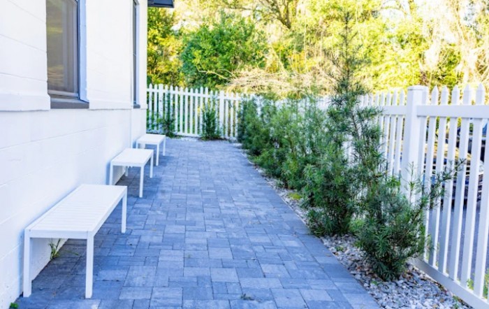 A paved pathway with benches alongside a white fence and shrubs.