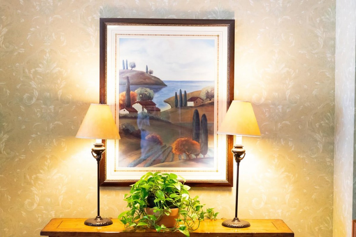 A cozy interior setting featuring a framed landscape painting above a console table, flanked by two elegant table lamps and adorned with a lush green plant.
