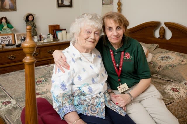 An elderly woman and her caregiver sit together on a bed, sharing a moment of companionship and affection.