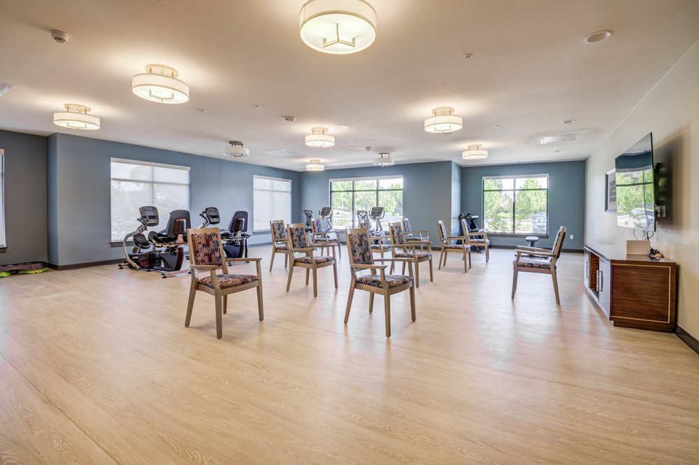 A spacious exercise room with chairs arranged in the center and stationary bikes along the wall, featuring large windows and wooden flooring.