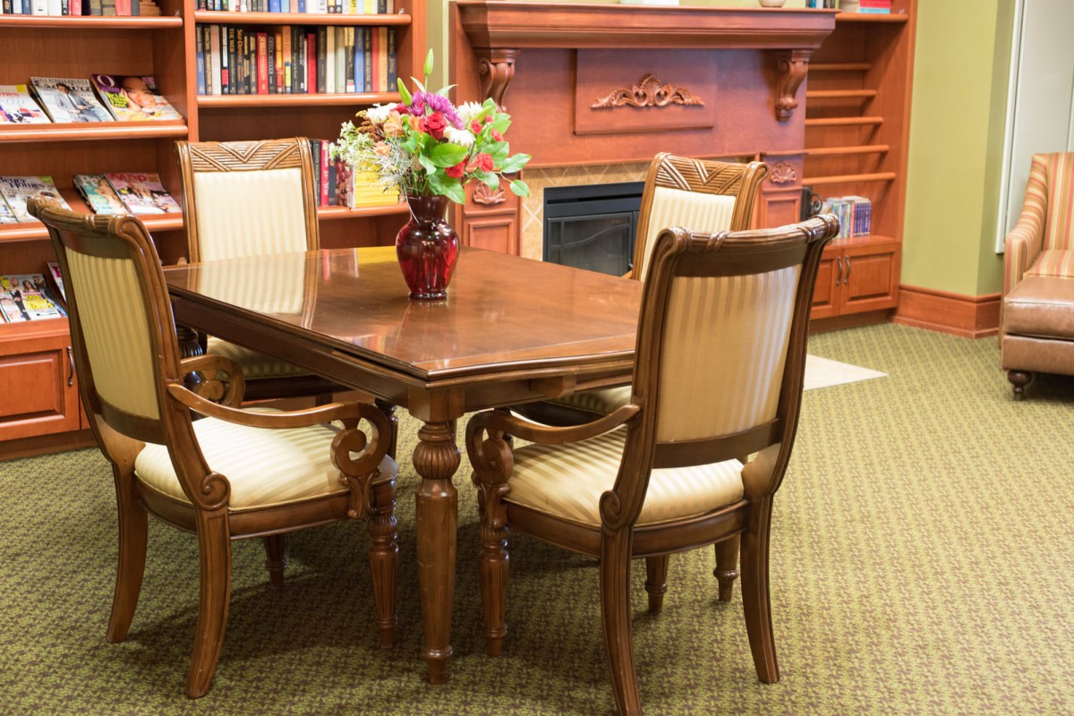 A cozy library setting with a wooden table and chairs, surrounded by a bookshelf and fireplace.