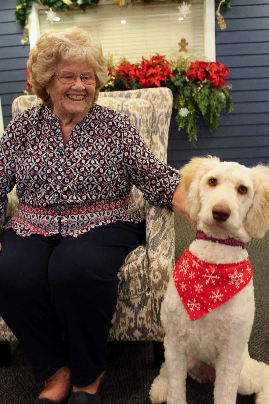 An elderly woman sitting on a chair indoors, smiling with a dog wearing a red bandana by her side.