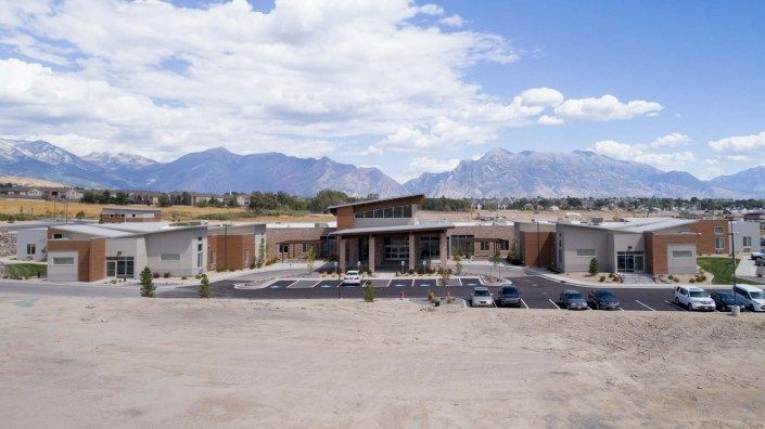 A modern building with a parking lot in front, set against a backdrop of mountains under a partly cloudy sky.