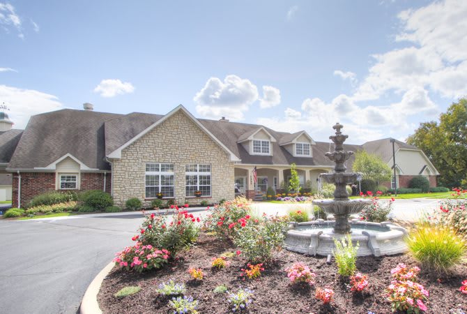 A building with a landscaped garden featuring colorful flowers and a fountain under a bright blue sky.