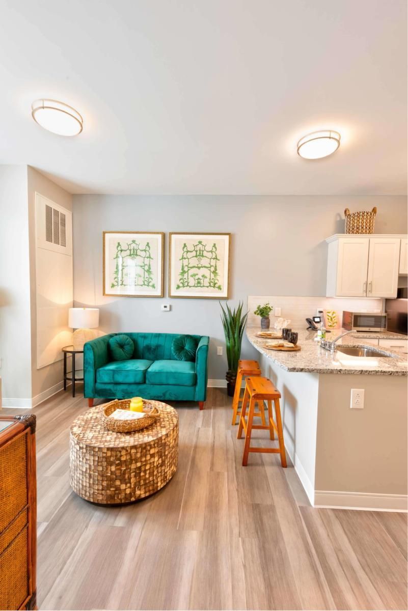 A modern living room with a cozy teal sofa, round coffee table, and an open kitchen area featuring bar stools and decorative plants.