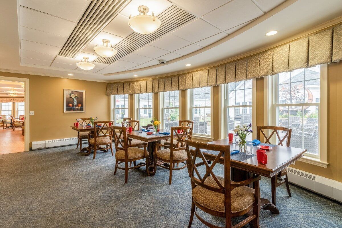 A cozy dining area with multiple tables and chairs, warmly lit and lined with large windows.