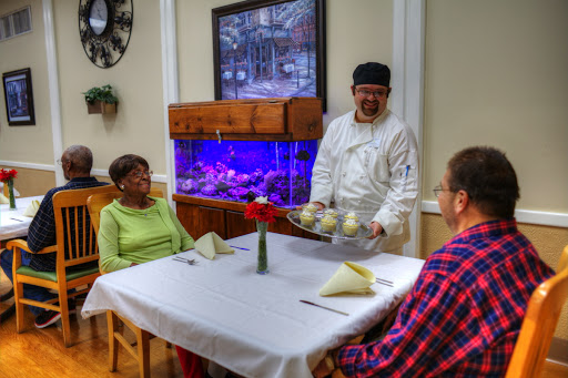 A chef serves a meal to two senior diners in a cozy dining room with an aquarium.