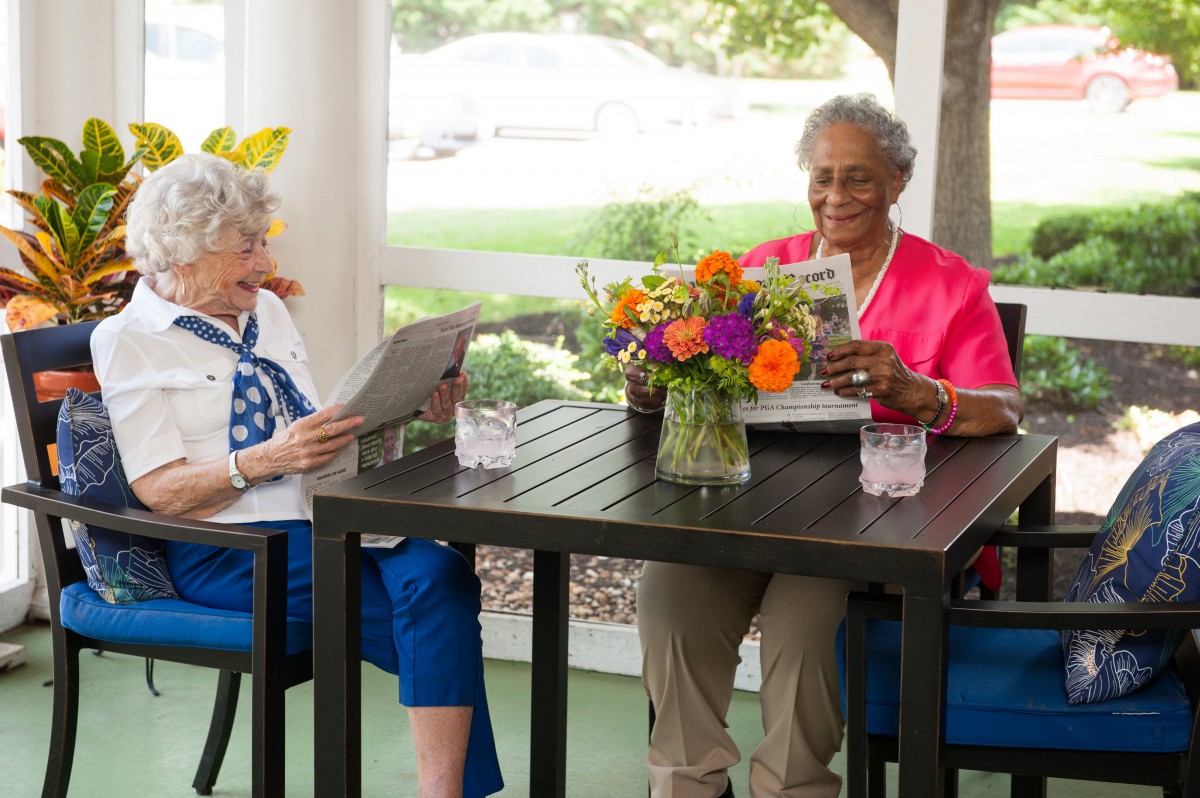 Two senior women enjoying a relaxed afternoon reading newspapers outdoors at a table with a colorful flower arrangement.
