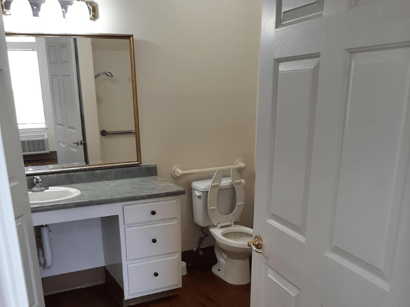 A bathroom featuring a sink, mirror, and toilet with white cabinets and an open door.