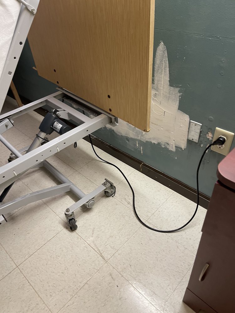 A hospital bed next to a power outlet, with a bedside table and medical equipment visible.