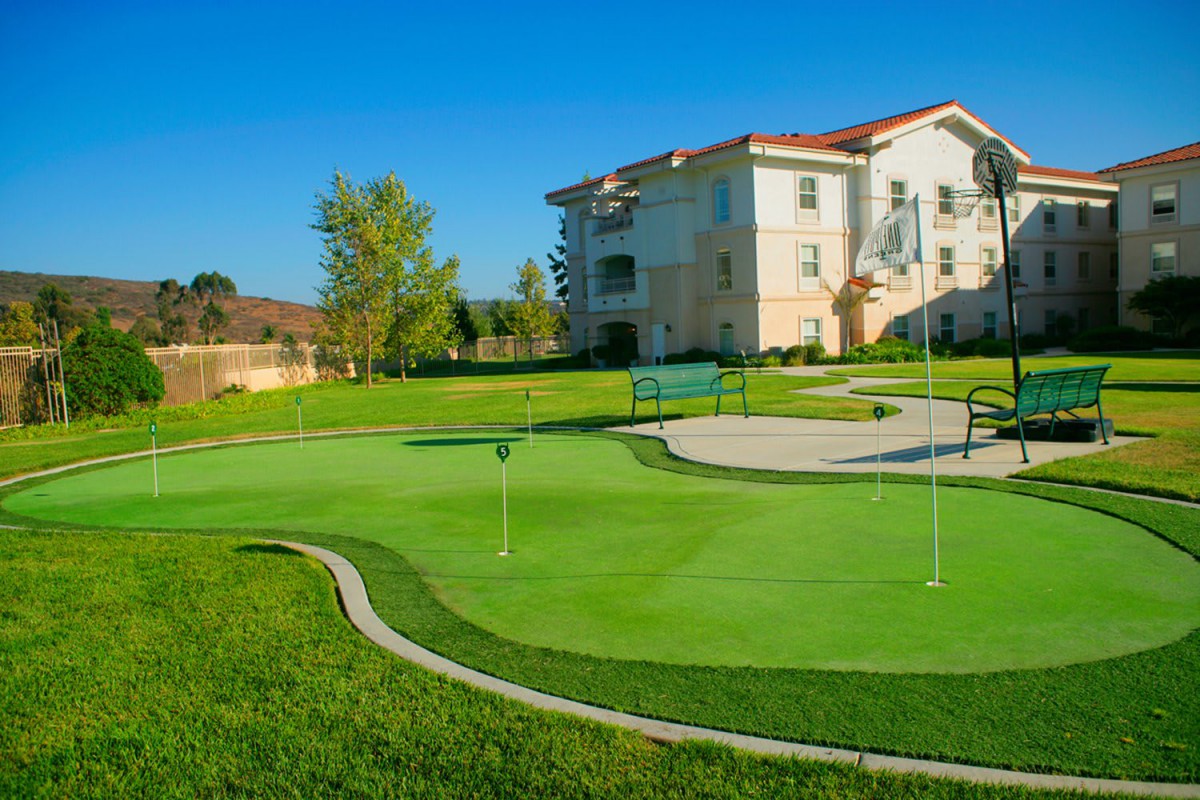 An outdoor putting green with a residential building in the background, surrounded by well-maintained grass and trees.