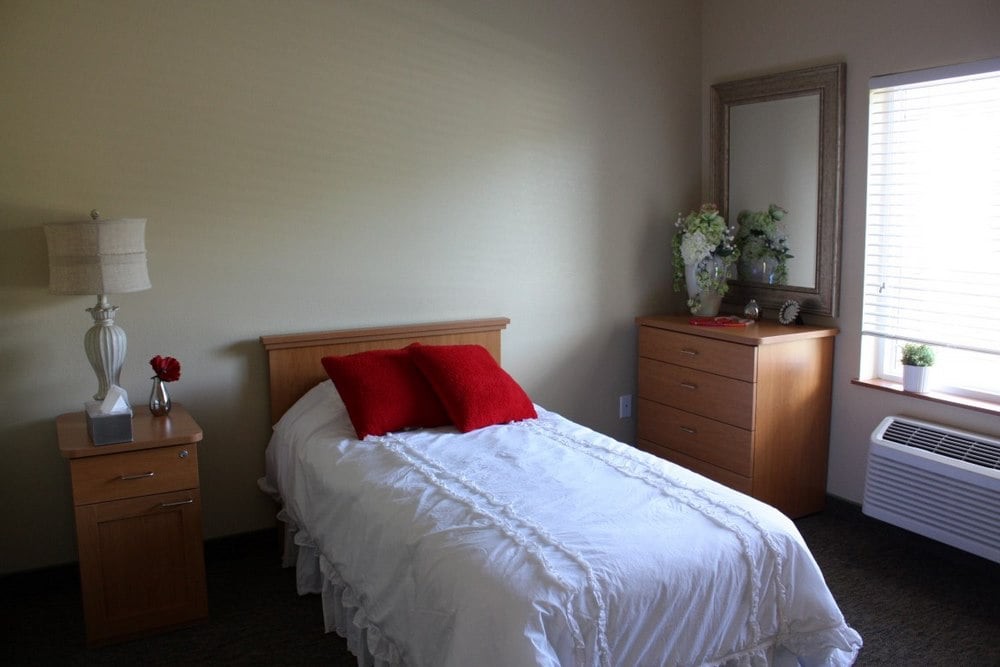 A minimalist bedroom with a single bed adorned with red pillows and white bedding, alongside a dresser and mirror.