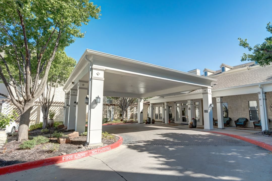 A view of a building entrance with a covered driveway and surrounding greenery on a sunny day.