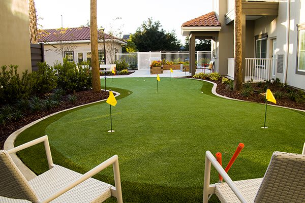 A backyard with a neatly maintained artificial turf putting green, surrounded by landscaping and patio furniture.