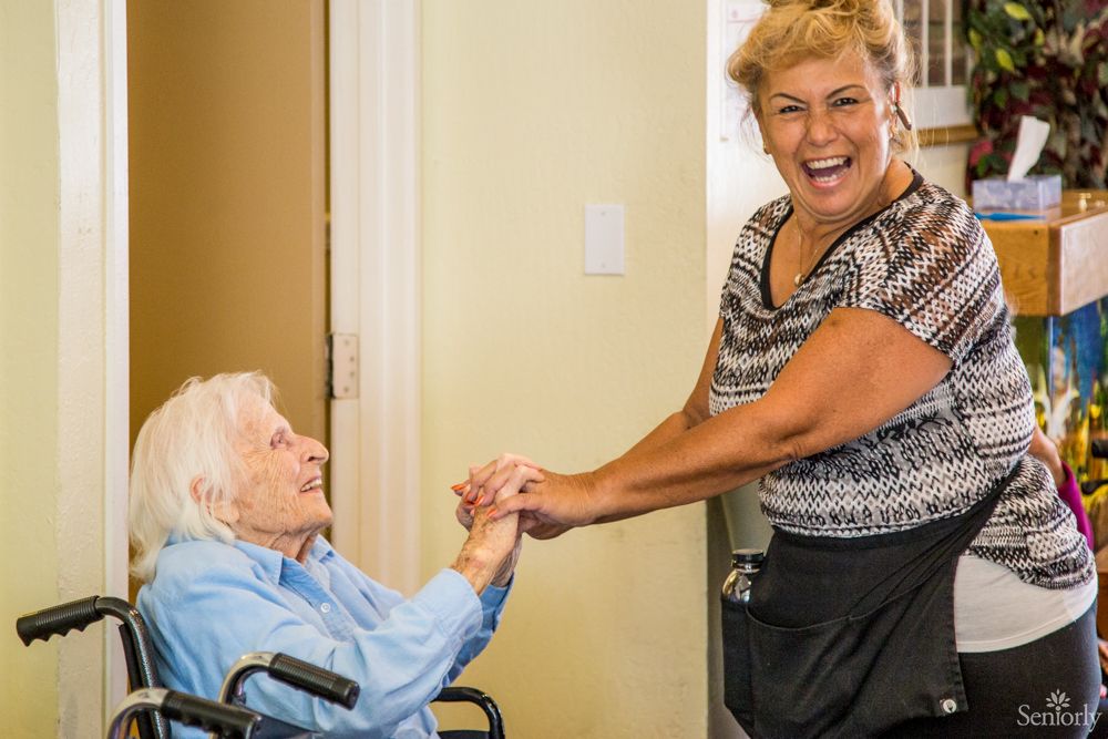 A smiling caregiver interacts warmly with an elderly woman in a wheelchair.