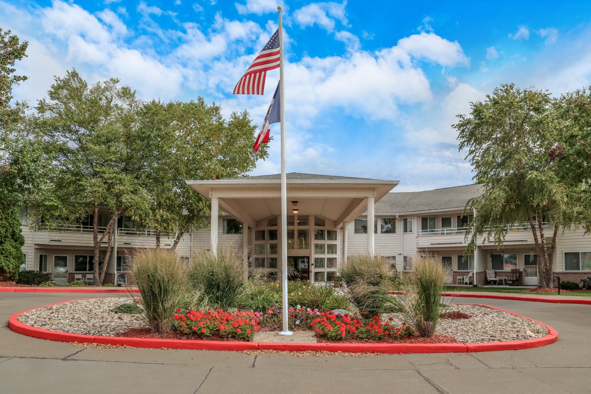 A well-maintained building entrance with flags and a colorful garden under a partly cloudy sky.