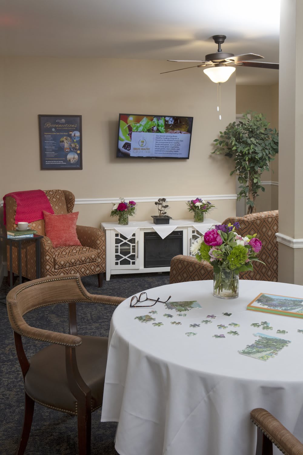A cozy living room area with a table decorated with flowers, comfortable chairs, and a television on the wall.
