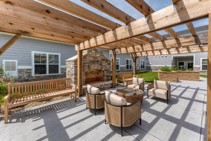 A cozy outdoor seating area under a wooden pergola, featuring comfortable chairs and a stone fireplace.