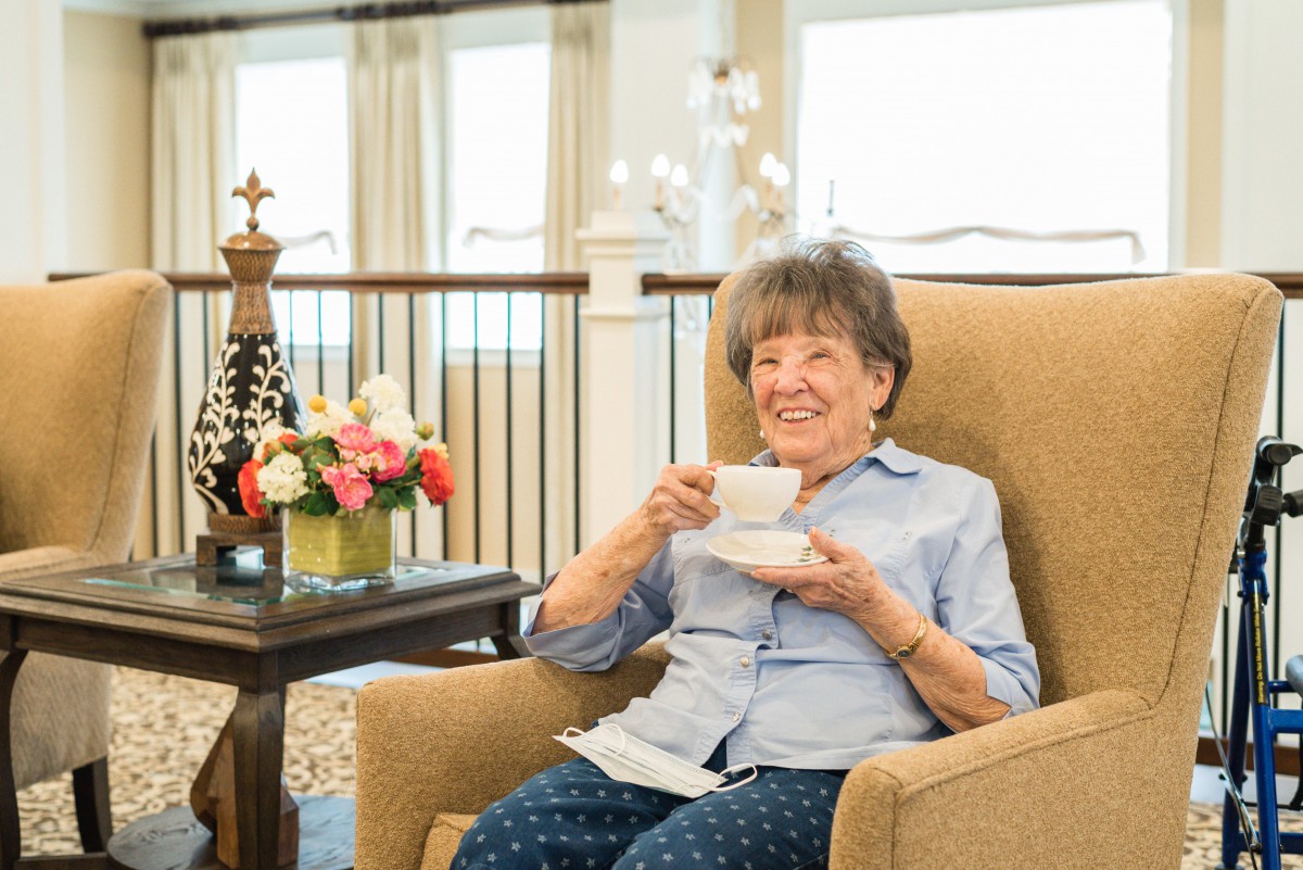 An elderly woman smiles while enjoying a cup of tea in an armchair, surrounded by a cozy indoor setting with flowers.
