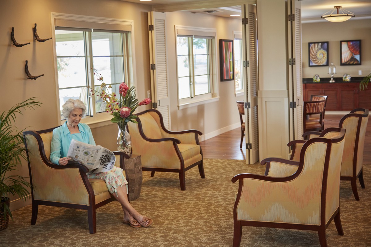An elderly woman reads a newspaper in a comfortable lounge area.
