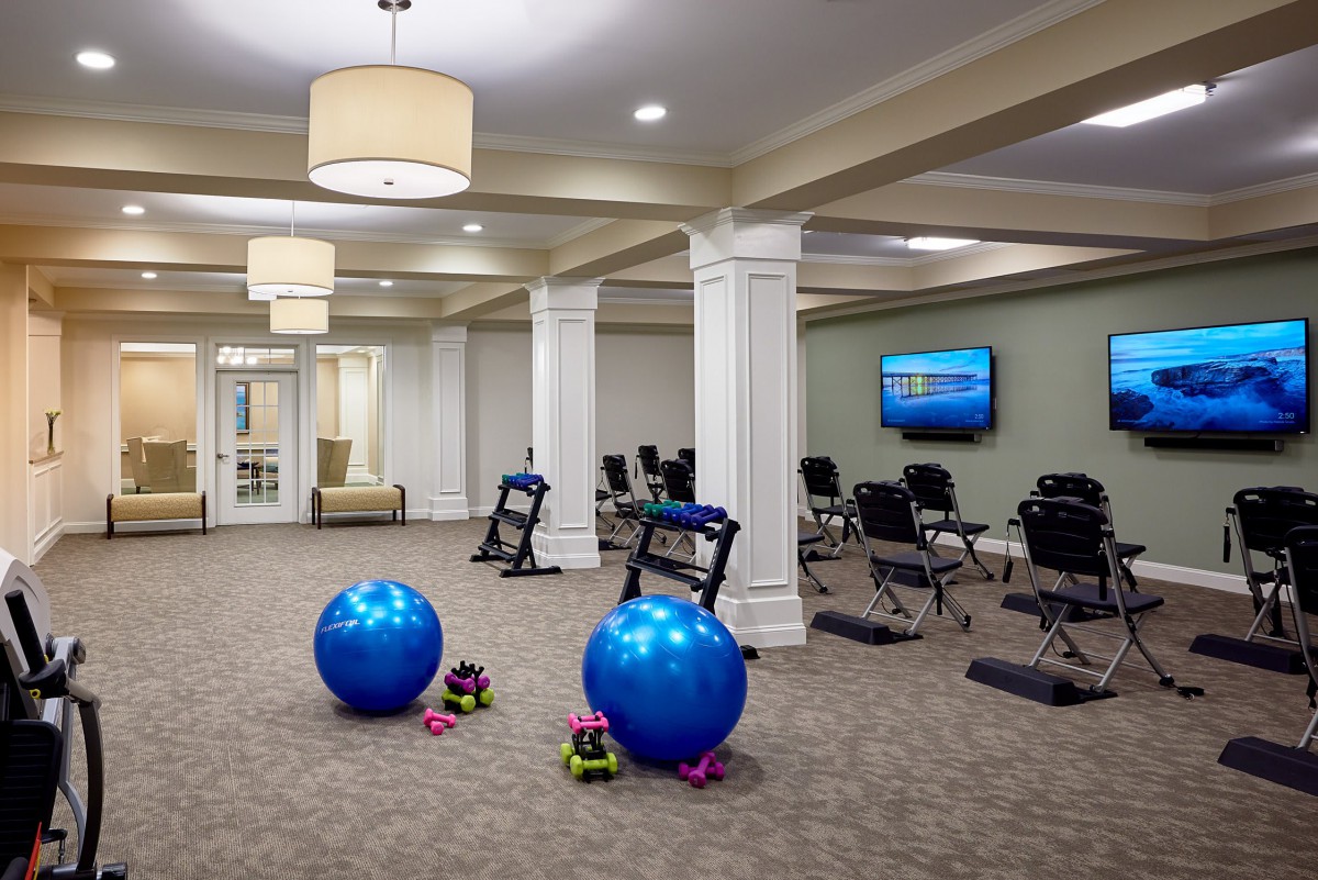 A spacious exercise room equipped with yoga balls, dumbbells, and chairs, featuring a carpeted floor and a television.