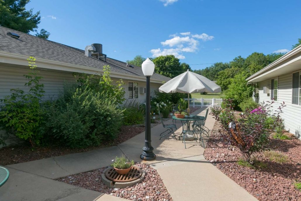 A sunny patio area with a table, chairs, and an umbrella surrounded by lush greenery.