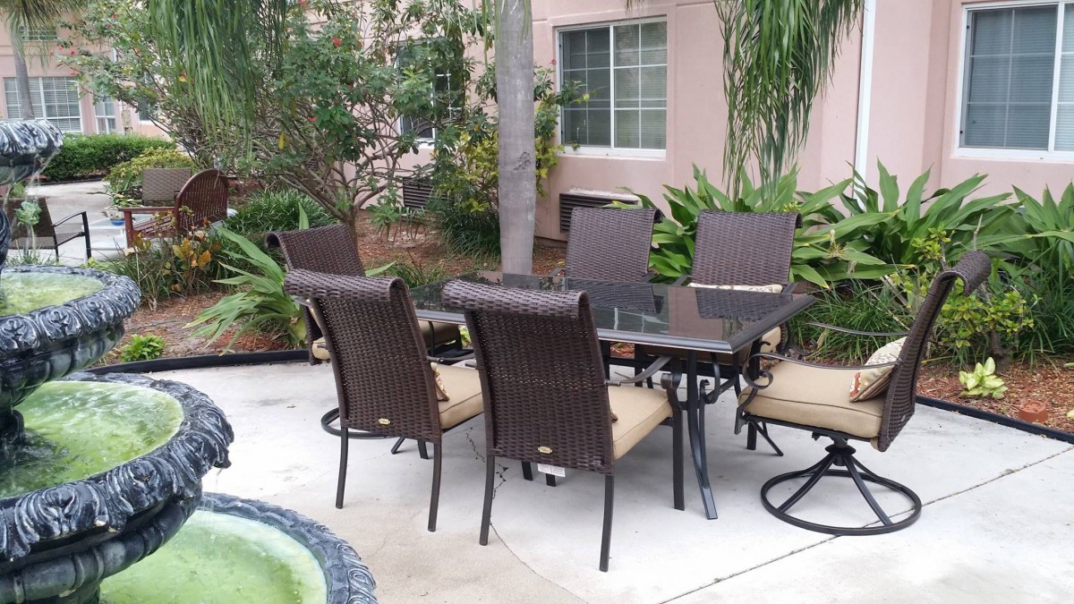 An outdoor patio set with a glass table and wicker chairs near a garden and pool.