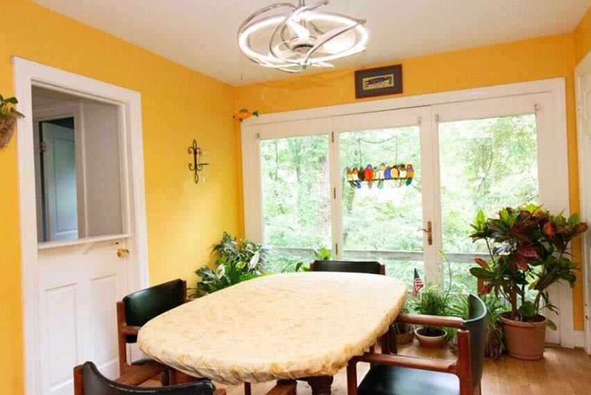 A bright dining room with yellow walls, featuring modern lighting, a table covered with a tablecloth, and several indoor plants by large windows.