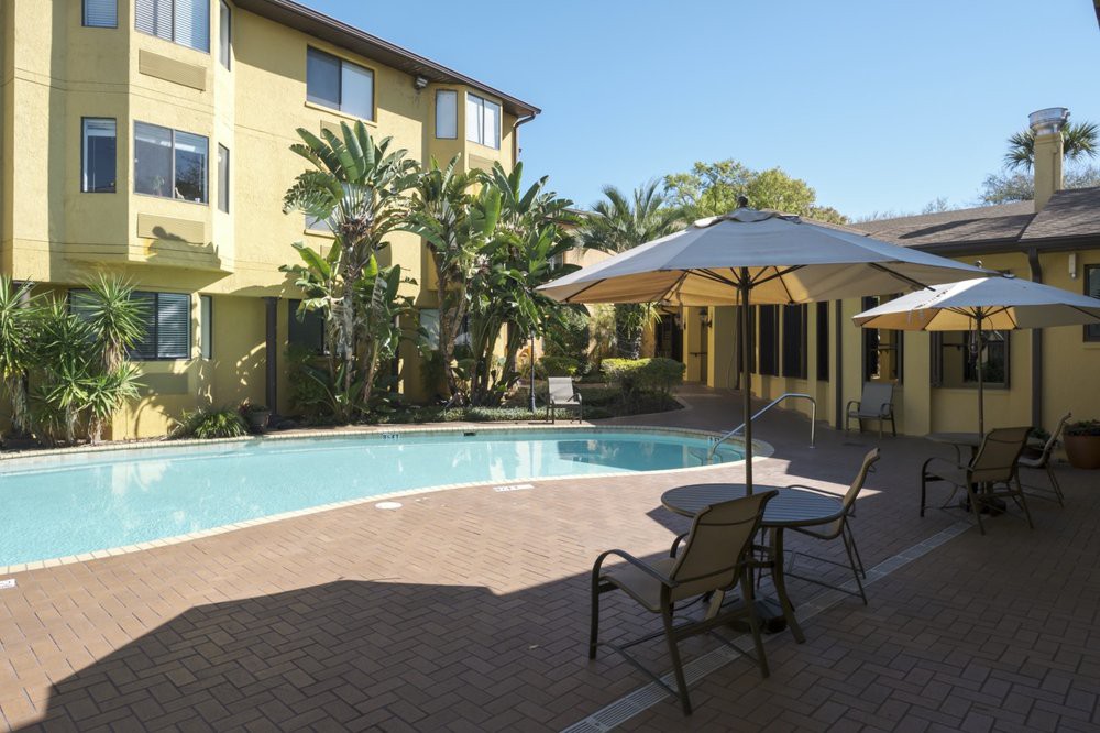 A serene outdoor swimming pool area with umbrellas and chairs, surrounded by tropical plants and an apartment building.
