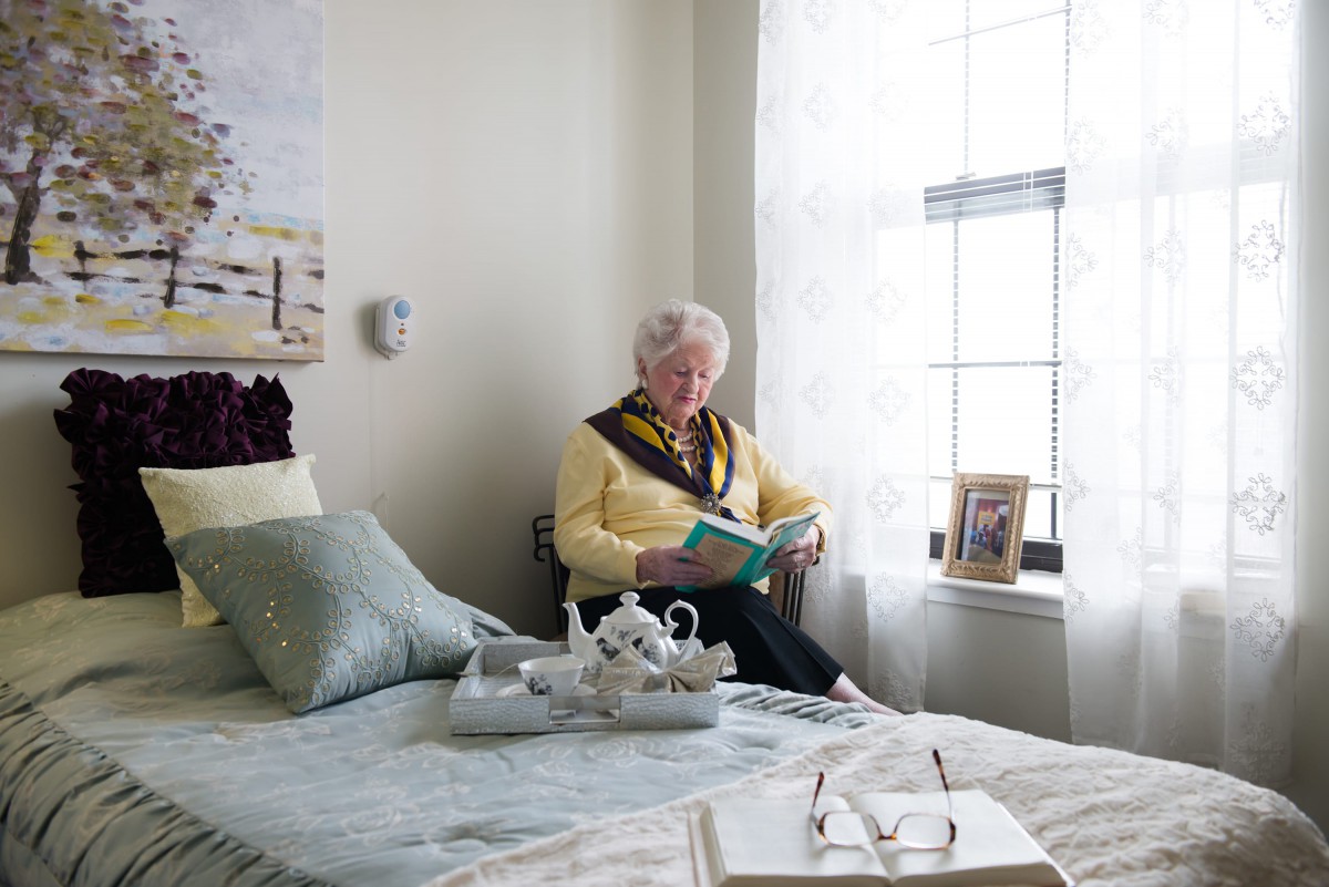 An elderly person sitting by the window, reading a book in a cozy bedroom with a tea set on the bed.