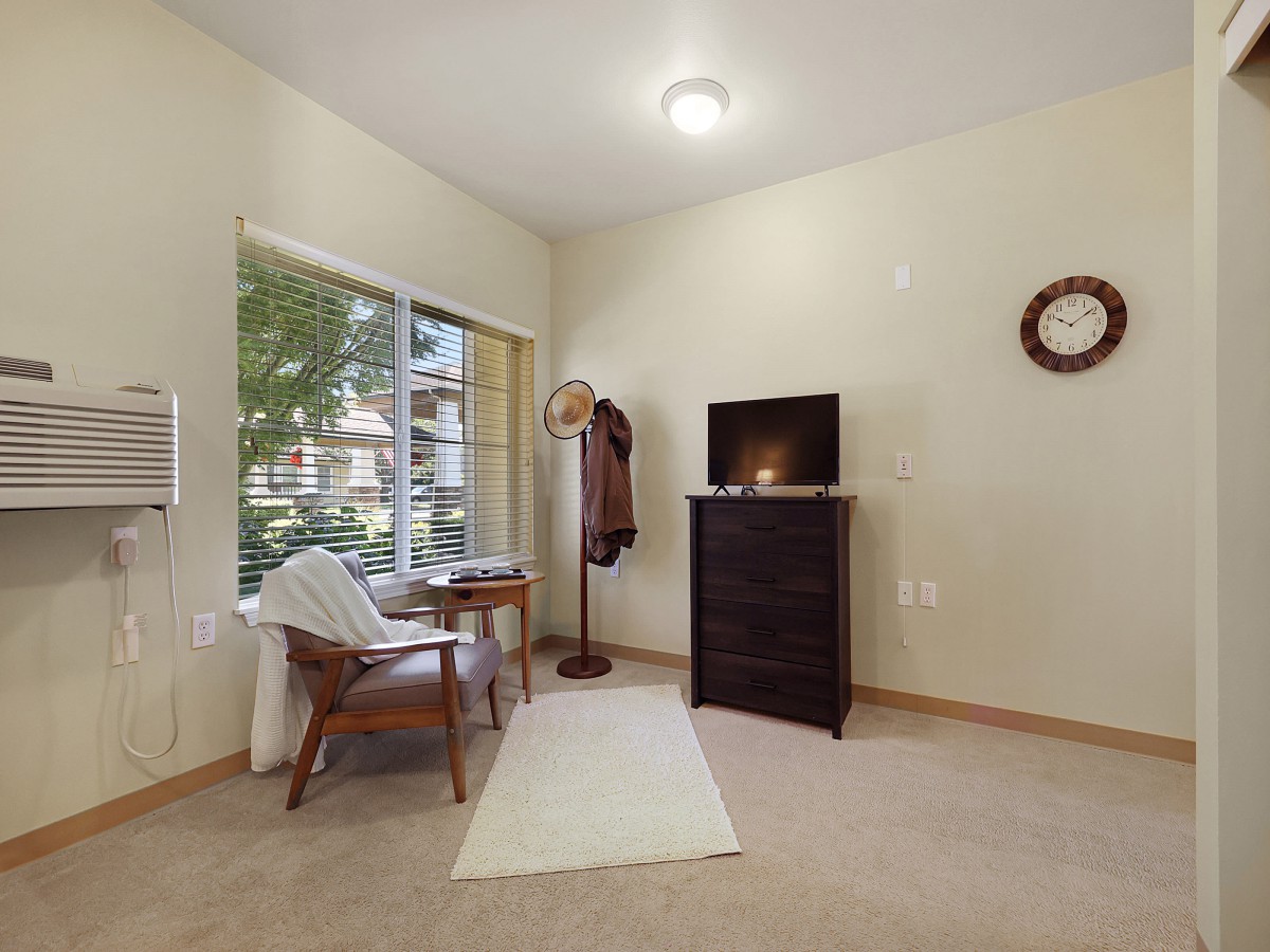 A cozy room with a chair and table by the window, a dresser with a television on top, and a wall clock.