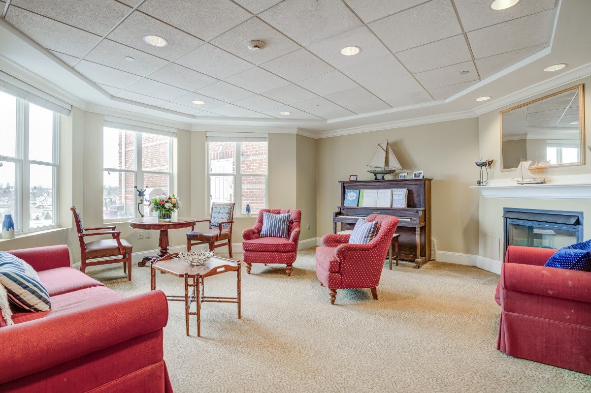 A cozy living room with red chairs and a piano, featuring large windows allowing natural light.