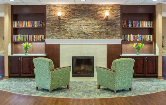 A cozy living room featuring a stone fireplace, two green armchairs, and bookshelves on either side.