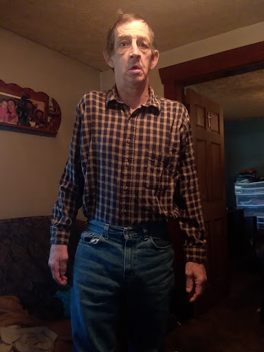 A man standing indoors wearing a checkered shirt and jeans.