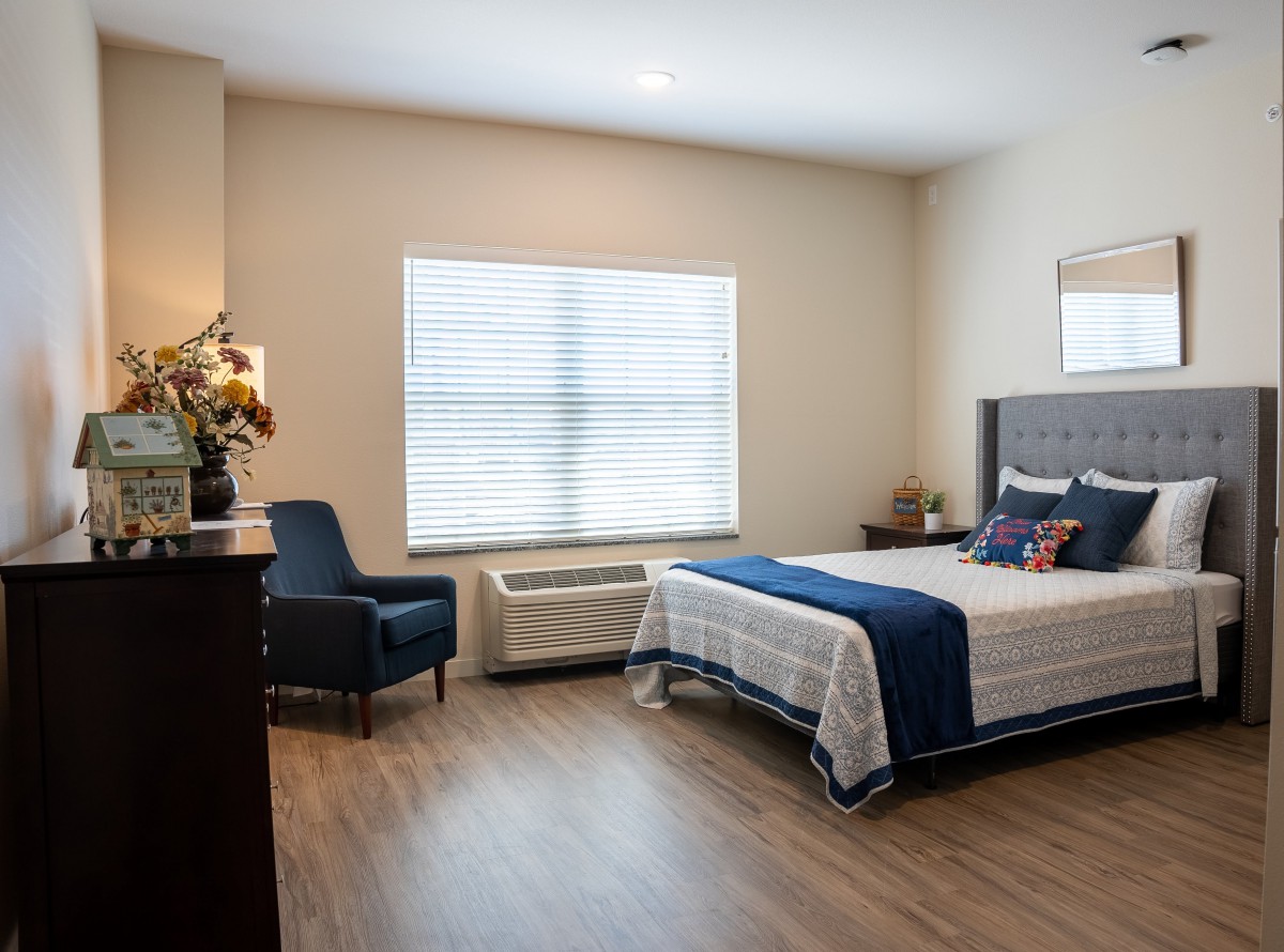 A modern bedroom with minimalist decor, featuring a bed, chair, and a large window providing natural light.