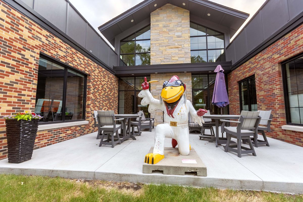 A bird mascot statue dressed in a white suit is displayed on the patio of a brick building with outdoor seating and a purple umbrella.