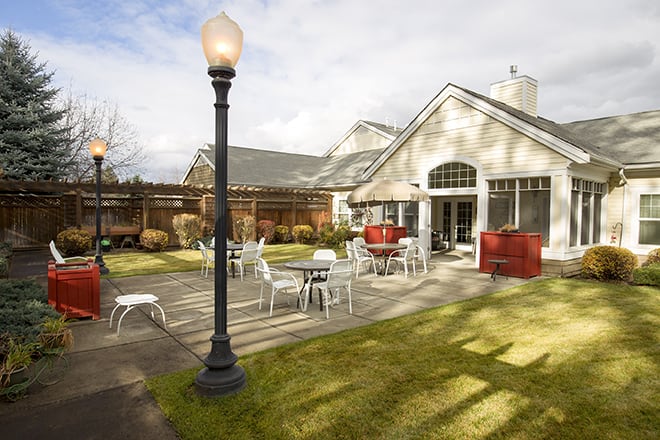 A cozy outdoor patio area with tables and chairs next to a building, featuring a lamp post and neatly maintained garden.
