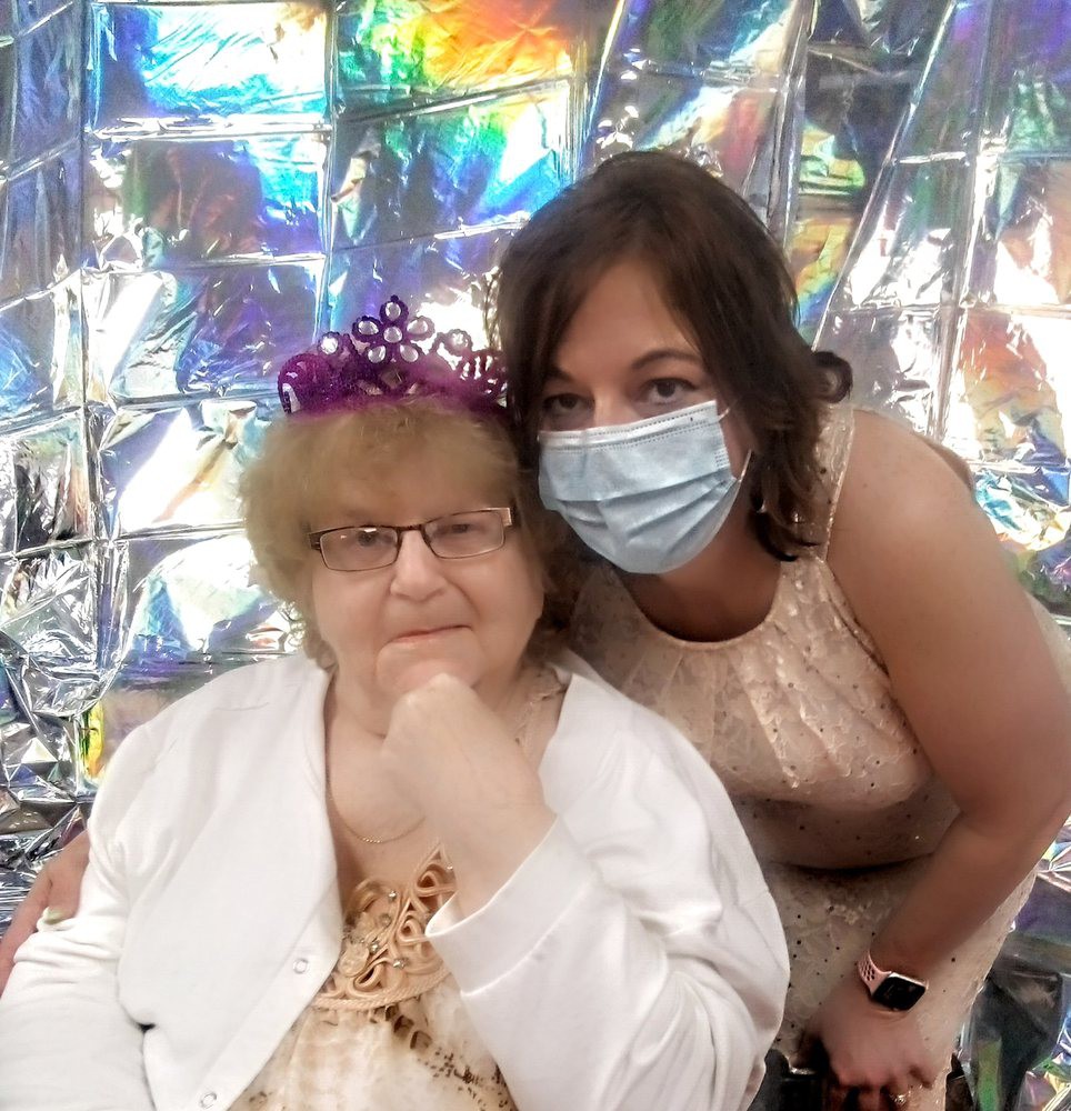 An elderly woman wearing a purple tiara sits beside another woman in a sparkling setting.