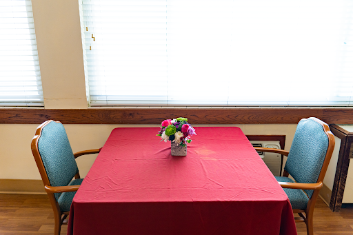 A neatly set table with a red tablecloth, accompanied by a small flower arrangement, beside a window.