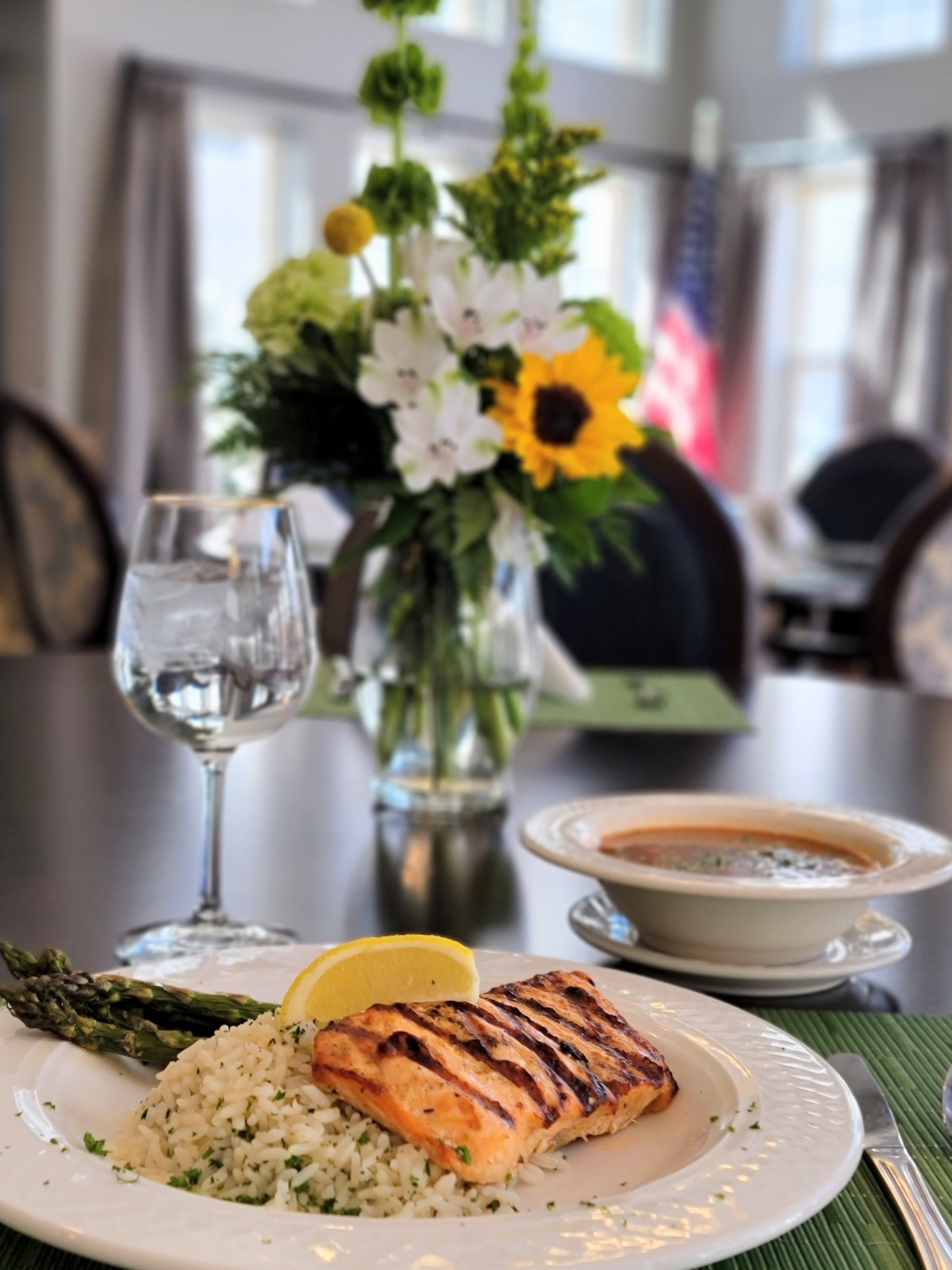 A beautifully arranged meal featuring grilled salmon with asparagus, rice, and a bowl of soup on a dining table adorned with a floral centerpiece.