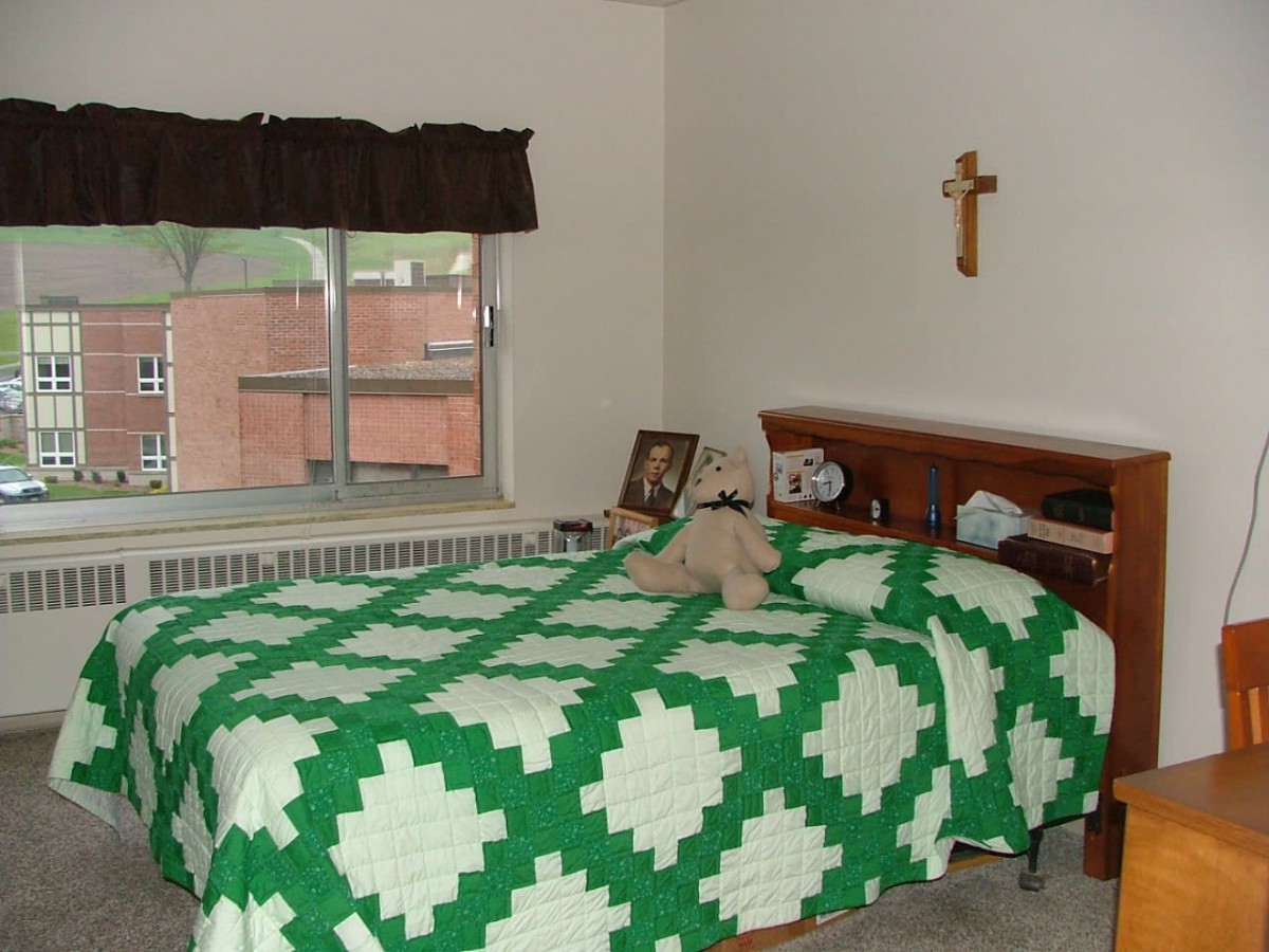 A cozy bedroom with a green and white quilt, a stuffed animal on the bed, and a cross hanging on the wall.